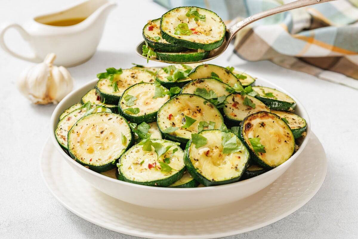 A bowl of sliced, roasted zucchini garnished with herbs, with a spoon serving a portion. A garlic bulb and a jug of sauce are in the background.