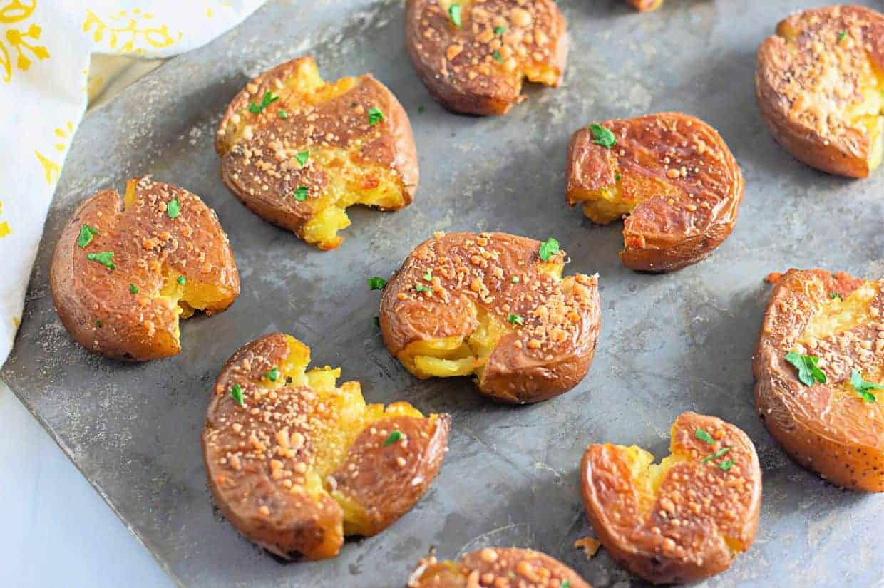Golden-brown smashed potatoes with crispy edges, sprinkled with Parmesan and fresh herbs, rest on a baking sheet. The melted cheese and garlic seasoning add a rich, savory touch.