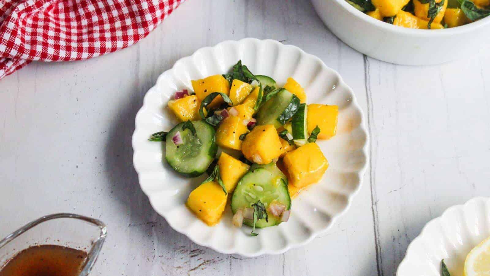 A white plate containing a salad with cucumber slices, mango chunks, red onion, and fresh herbs sits on a white surface.