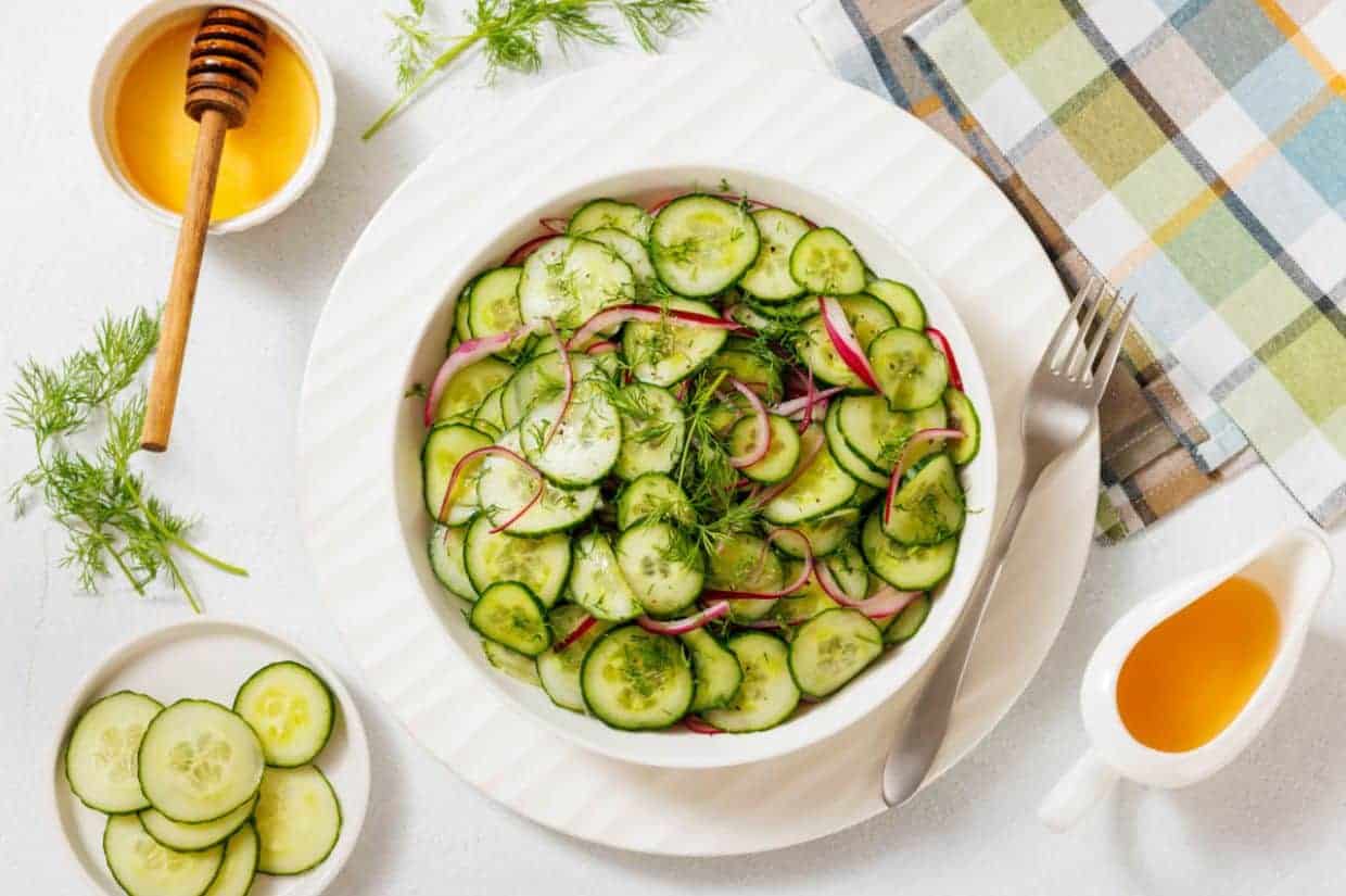 A fresh cucumber and red onion salad garnished with dill in a white bowl, accompanied by a honey dipper, a small dish of sliced cucumbers, a plaid napkin, and a jug of vinaigrette on a white table.