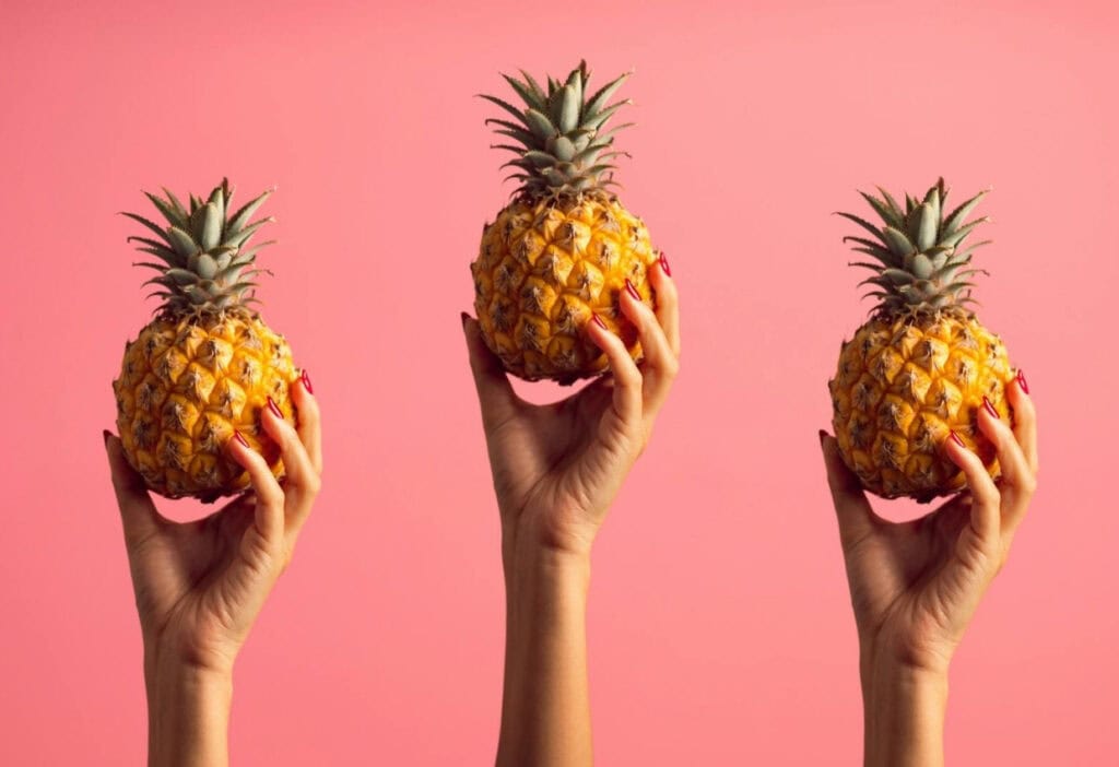 Three hands each hold a pineapple against a solid pink background.