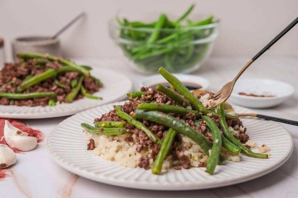 A plate of cooked ground beef and green beans served over rice, with chopsticks picking up a portion. A bowl of green beans and small dishes of sauces are in the background.