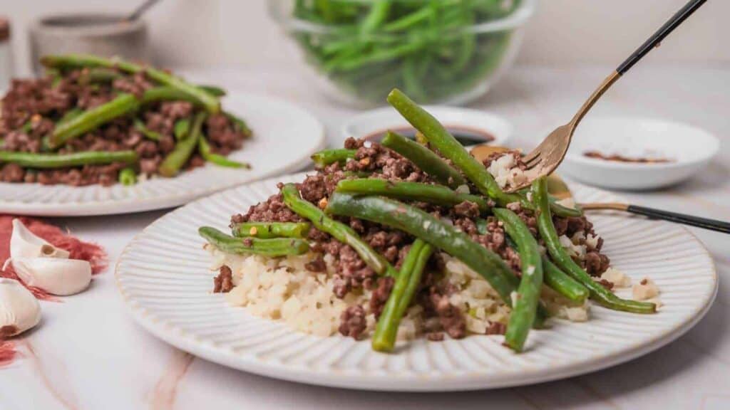 A plate of ground beef and green beans served over rice, with a fork resting on the plate. Another plate and bowl of green beans are in the background.