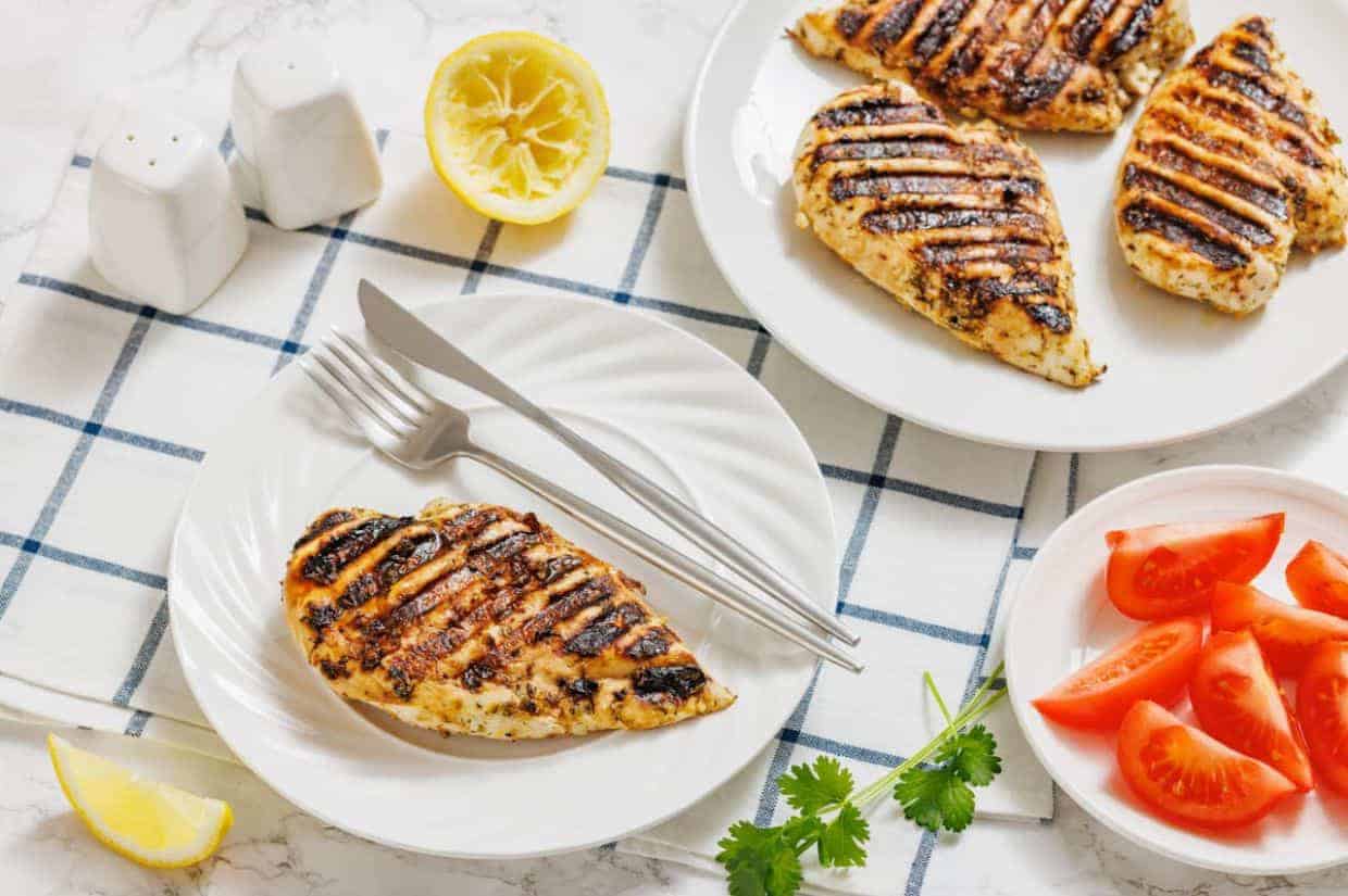 Grilled chicken breast on a white plate with fork and knife, sliced tomatoes, lemon halves, cilantro, and salt and pepper shakers on a checkered cloth.