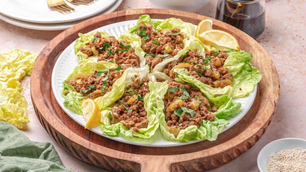 A plate of lettuce wraps filled with minced meat and vegetables, garnished with herbs and served with lemon wedges.