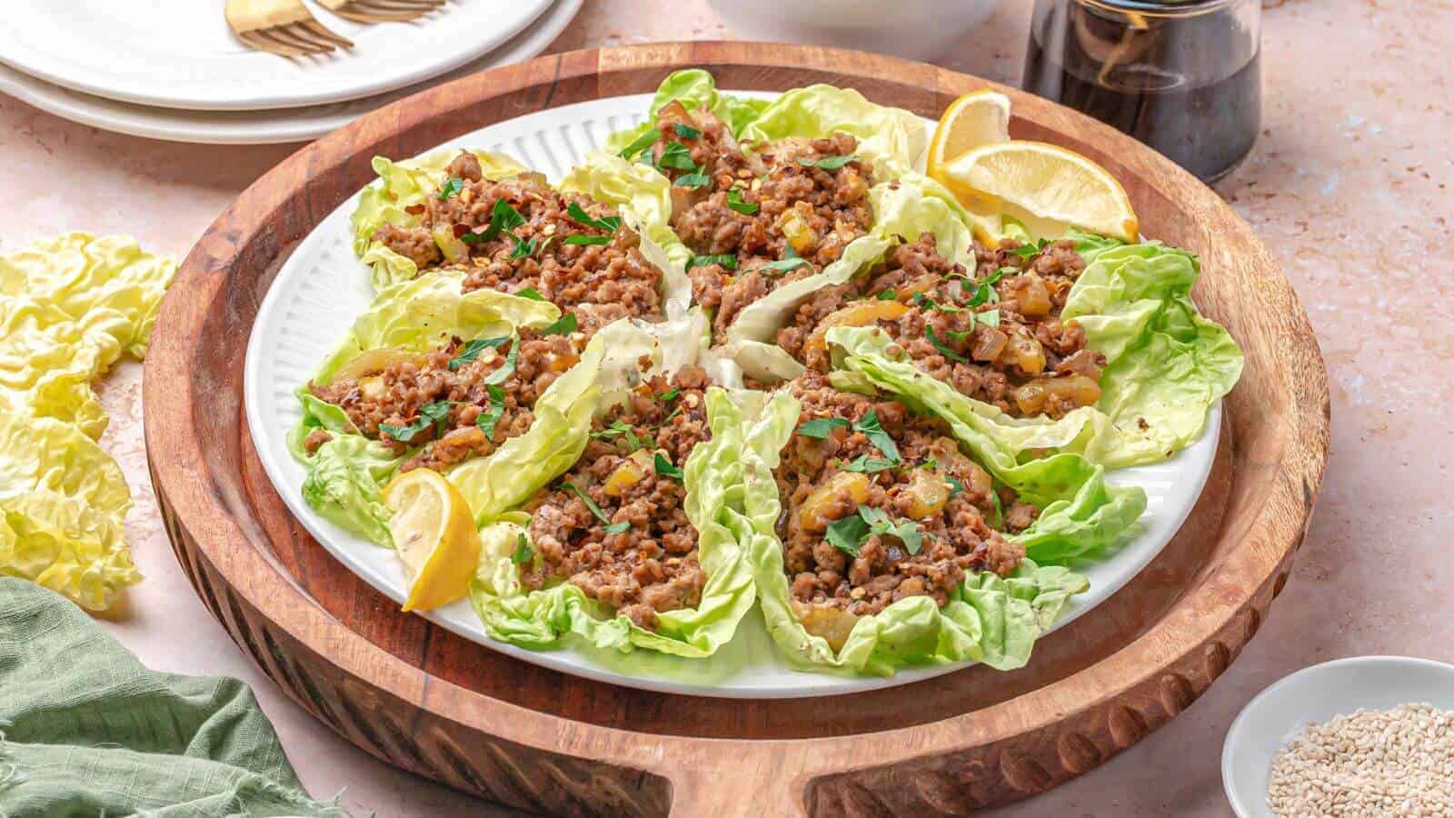 A plate of lettuce wraps filled with seasoned minced meat and garnished with herbs, served with lemon wedges on a wooden tray.