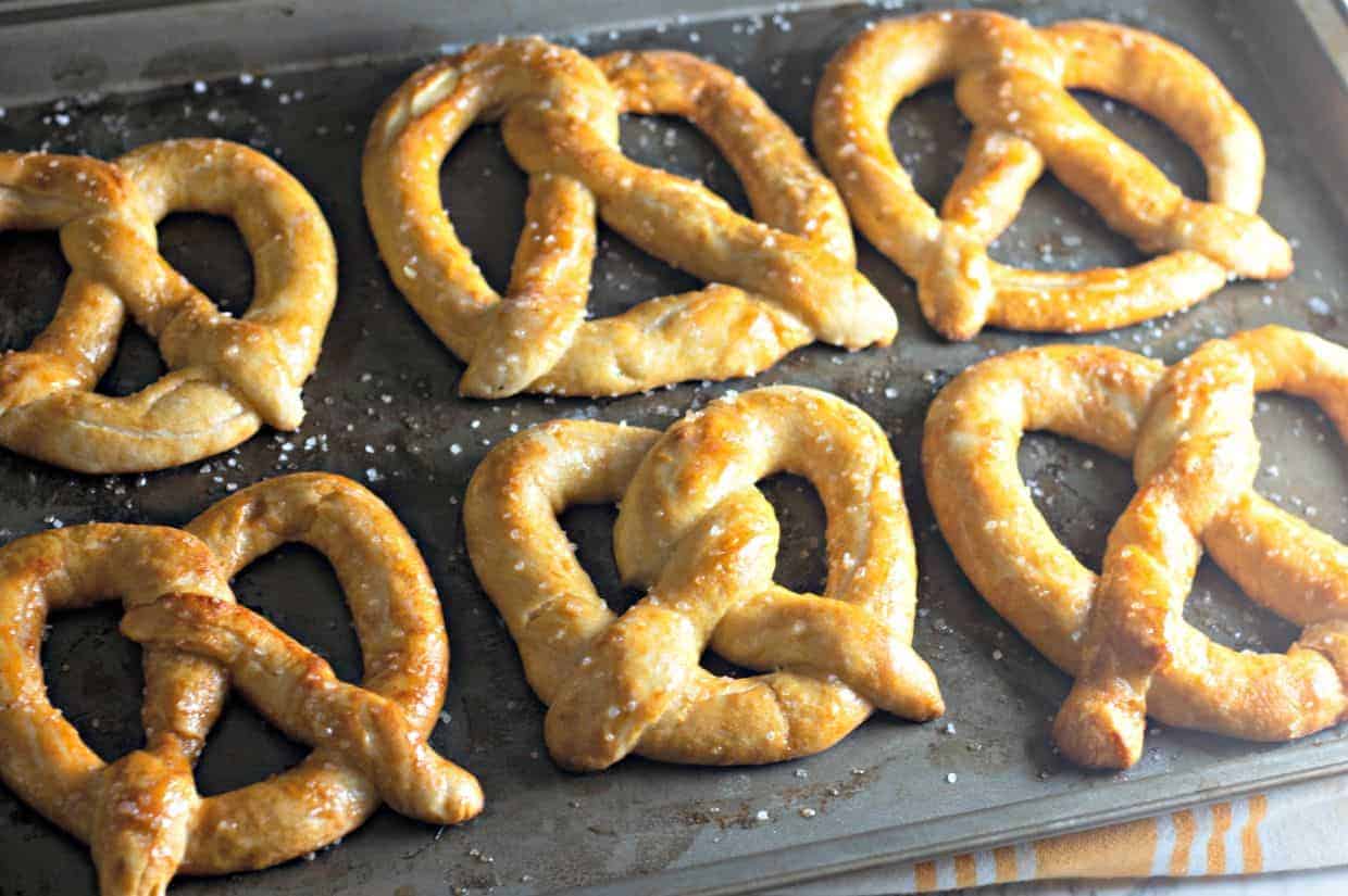 A baking sheet filled with six golden-brown homemade soft pretzels, sprinkled with coarse salt.