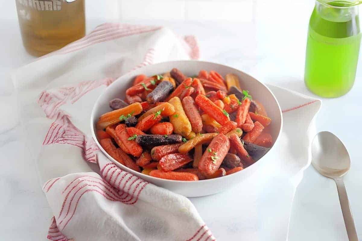 A white bowl filled with honey-roasted baby carrots sits on a white cloth with red stripes, next to a spoon and a green bottle on a light surface.
