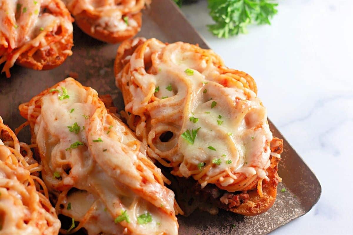 Toasted bread slices topped with spaghetti, meat sauce, melted cheese, and garnished with chopped parsley on a metal baking sheet.