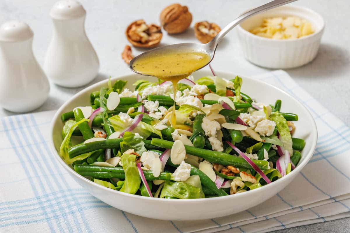 A spoon pours dressing over a salad with green beans, feta cheese, red onion, almonds, and walnuts in a white bowl on a checkered cloth. Salt, pepper, and parmesan are in the background.