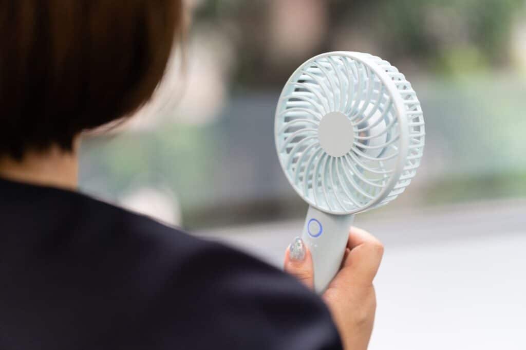 A person holds a small, handheld electric fan&mdash;one of those stylish coolers&mdash;facing away from the camera. The background appears blurred and out of focus.