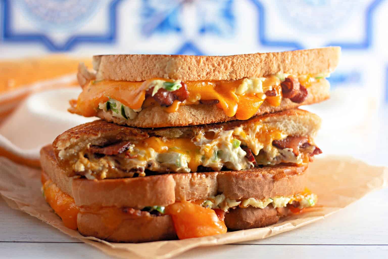 Two Jalapeño Popper Grilled Cheese sandwiches with melted cheddar, bacon, and a creamy filling are stacked on parchment paper, with cheese oozing out from the sides.
