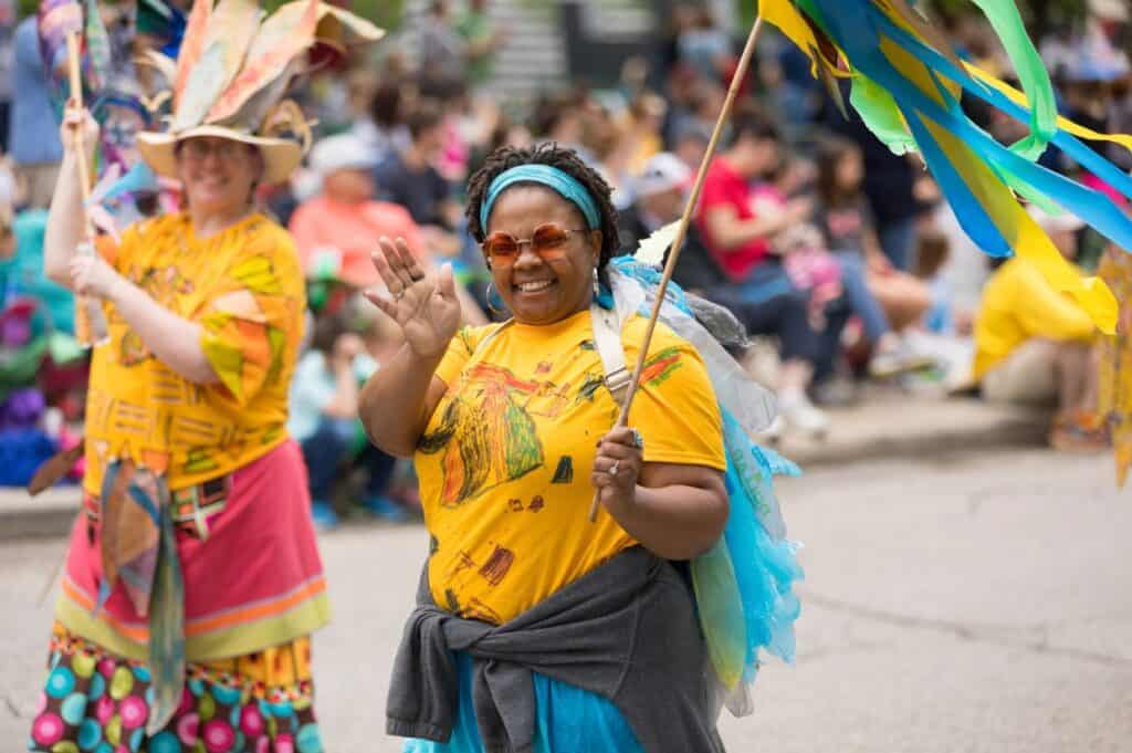 Two people in bright costumes walk in a Juneteenth parade; one woman smiles and waves at the camera while holding a colorful flag, with a crowd seated in the background.