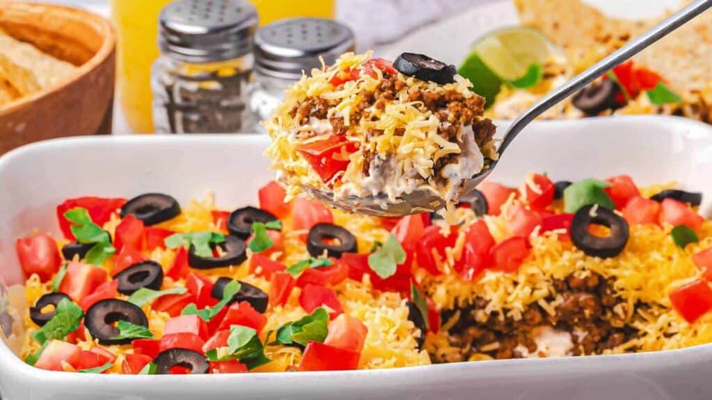 A spoon lifts a serving of layered taco dip topped with shredded cheese, tomatoes, black olives, and herbs from a white dish.
