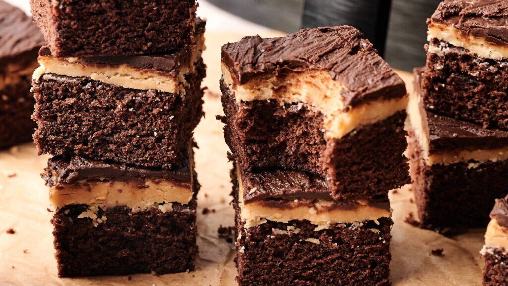 Stacked chocolate brownies with a peanut butter layer and chocolate frosting, displayed on a wooden surface.