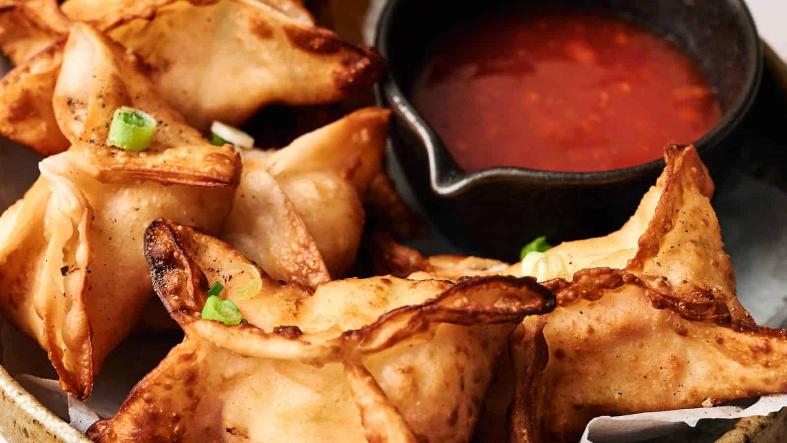 Crispy wontons garnished with green onions are served next to a small black bowl of red dipping sauce.