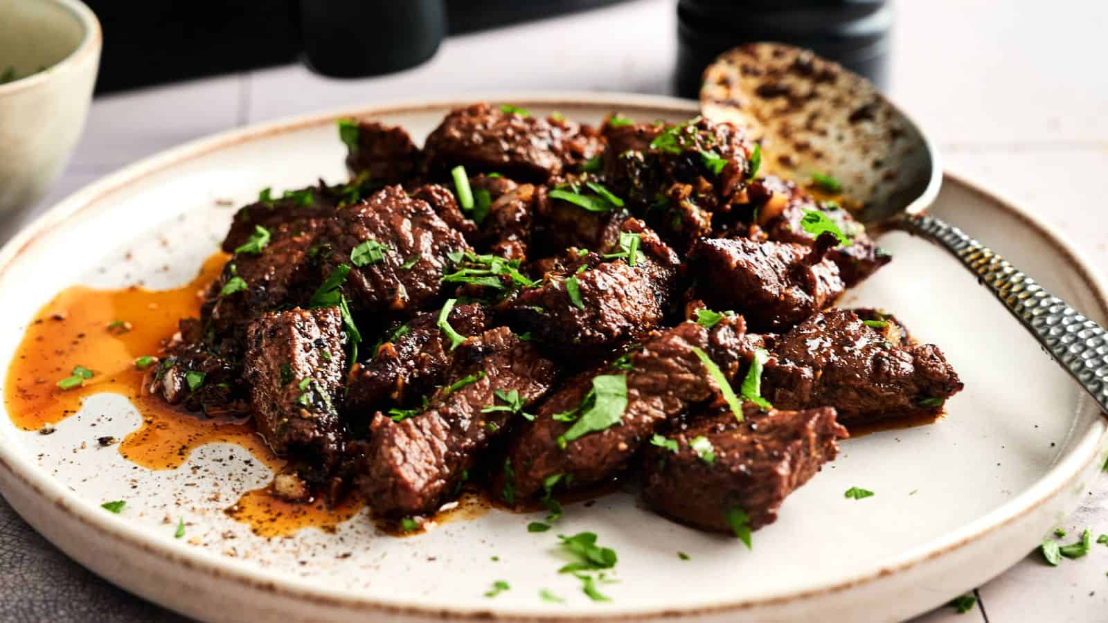 A plate of cooked beef chunks garnished with chopped parsley, with a spoon resting on the side and some sauce visible on the plate.
