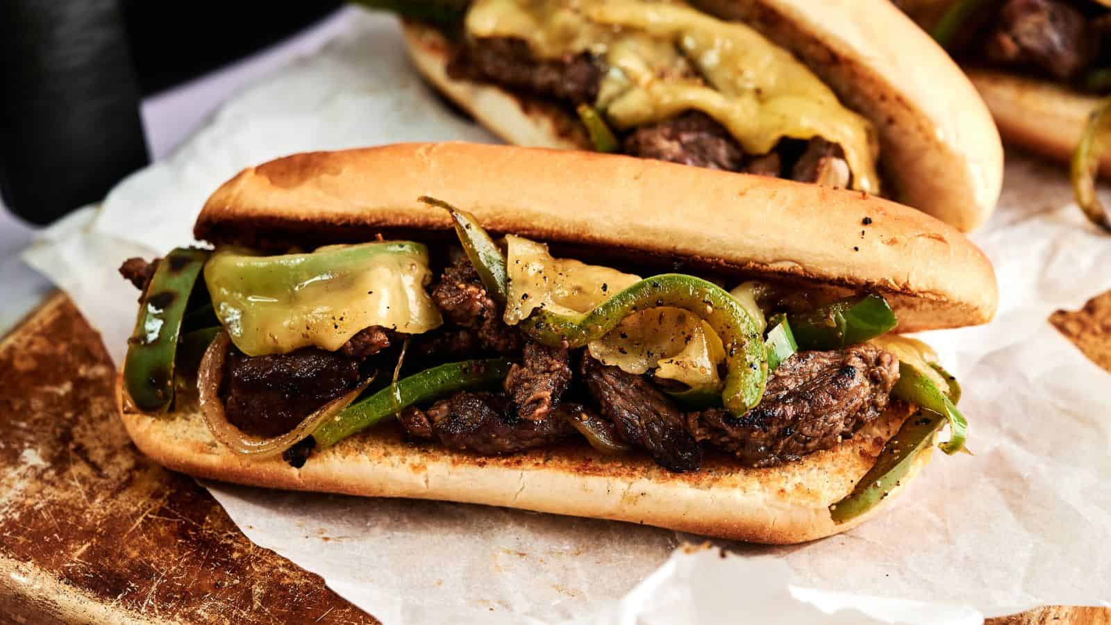 A sandwich roll filled with sliced steak, melted cheese, green bell peppers, and onions sits on parchment paper atop a wooden surface.