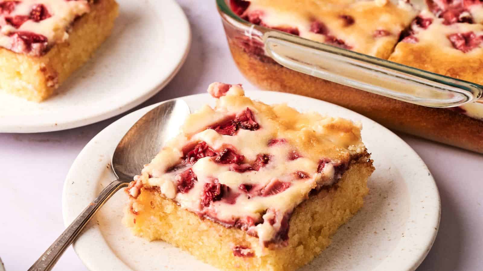 A square slice of strawberry-topped cake on a plate with a spoon, with more cake in a glass baking dish in the background.