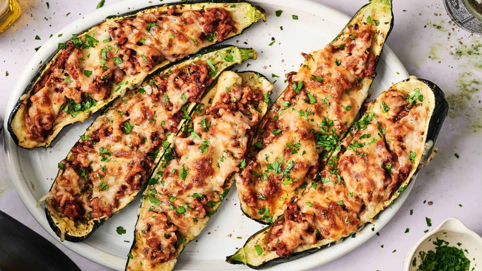 Five zucchini boats filled with a minced meat sauce and topped with melted cheese, garnished with chopped herbs, arranged on a white oval plate.