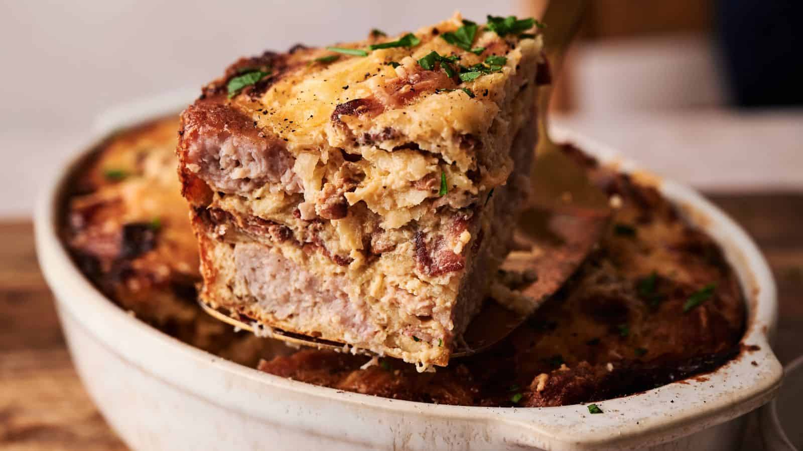 A close-up of a serving of savory bread pudding with bacon and herbs being lifted from a white baking dish.
