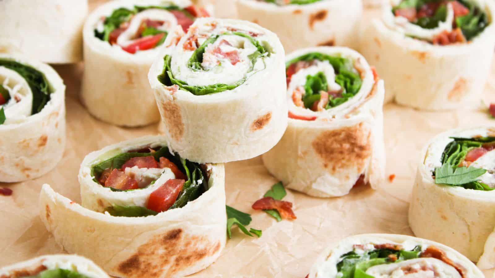 Several tortilla pinwheels filled with leafy greens, tomato, and possibly cheese or meat, arranged on parchment paper.