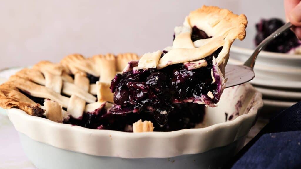 A slice of blueberry pie with a lattice crust is being lifted from a ceramic pie dish, revealing a juicy berry filling.
