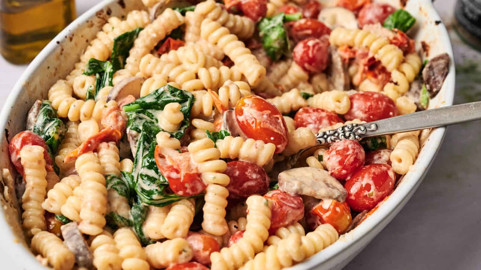 A white dish filled with cooked fusilli pasta, cherry tomatoes, spinach, and mushrooms in a creamy sauce, with a silver fork resting inside.