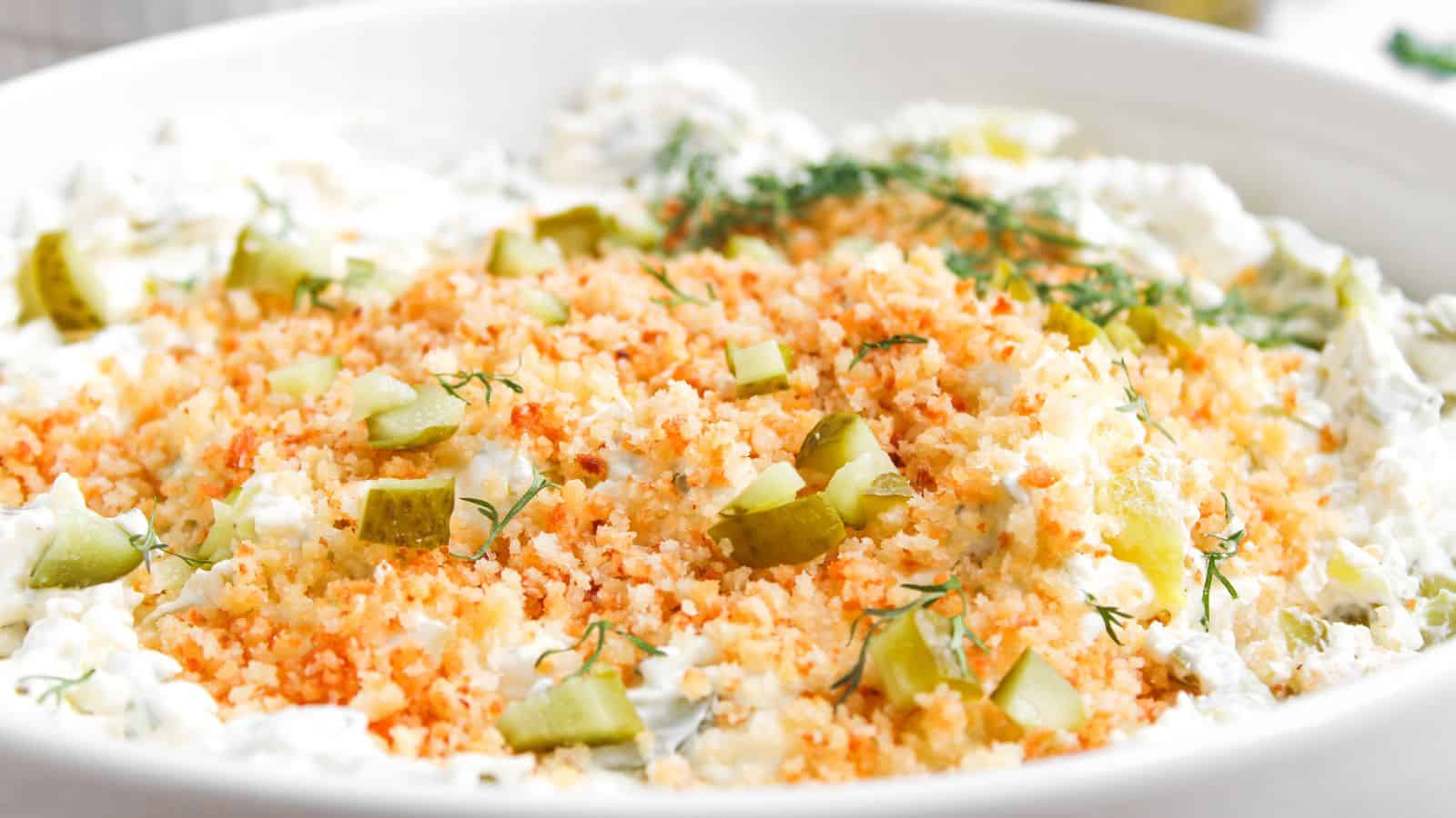 A close-up of a creamy dip topped with toasted breadcrumbs, chopped pickles, and fresh dill in a white bowl.