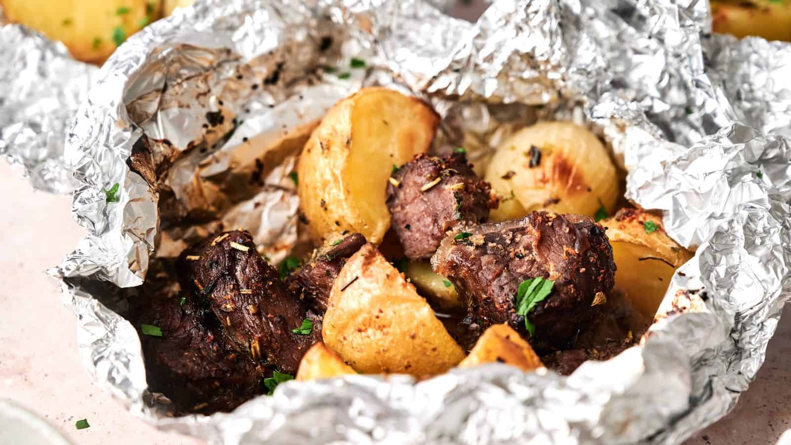 Chunks of cooked beef and roasted potatoes served in an open aluminum foil packet, garnished with herbs.