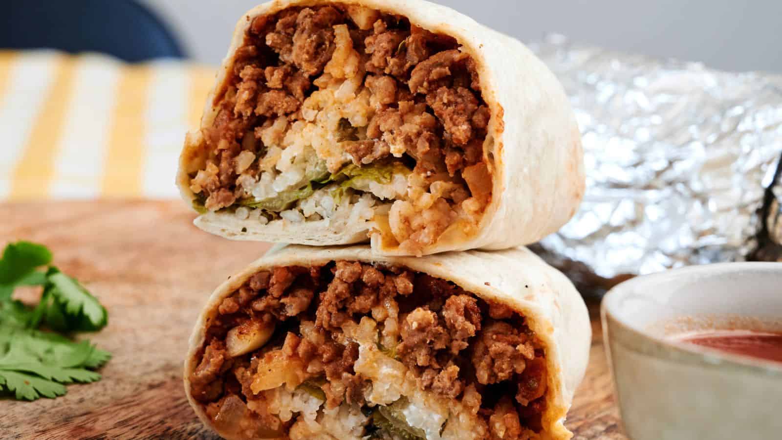 Three foil-wrapped burrito halves filled with ground meat, cheese, lettuce, and tomato are stacked on a pink plate.