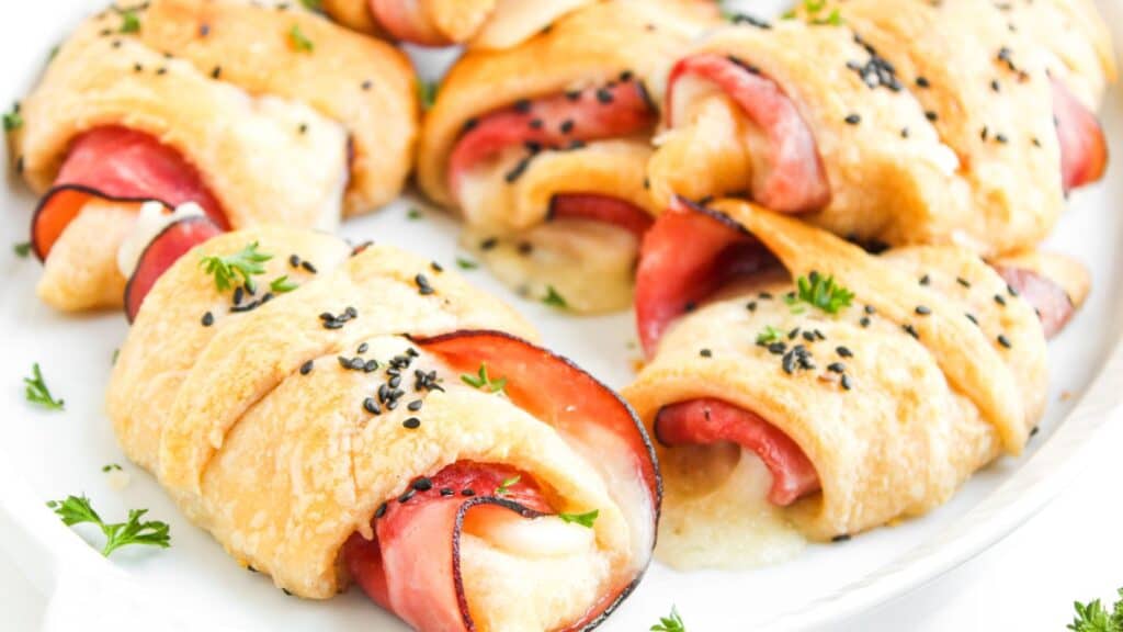 A plate of baked crescent rolls filled with ham and melted cheese, topped with sesame seeds and garnished with chopped parsley.