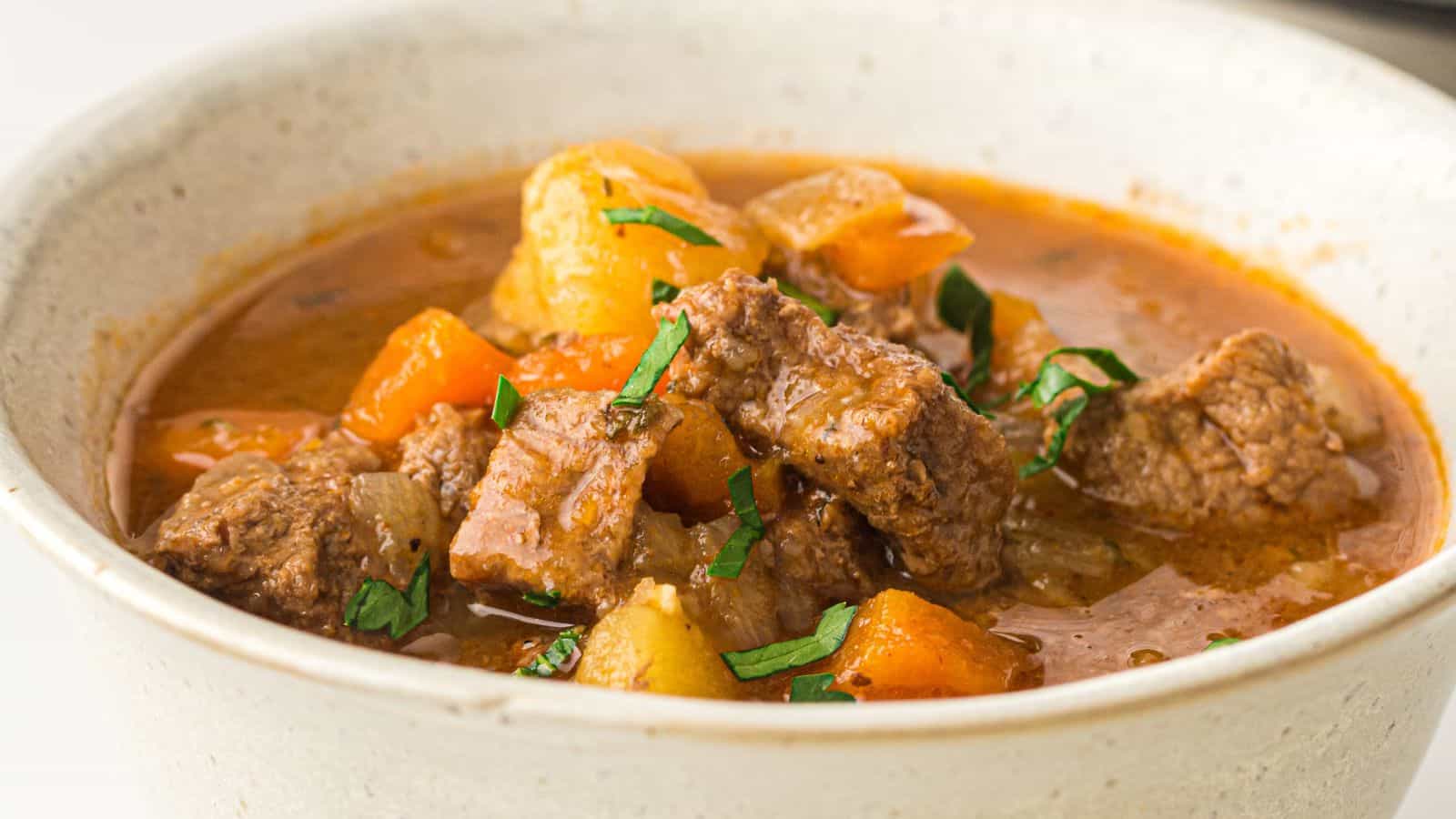 A bowl of beef stew with chunks of beef, potatoes, and carrots in a reddish broth, garnished with chopped herbs.