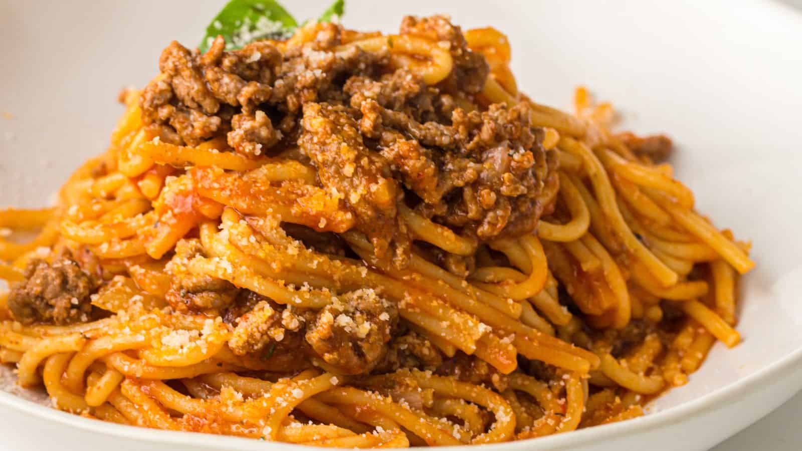 A close-up of spaghetti topped with ground meat and tomato sauce, garnished with grated cheese and a basil leaf, served on a white plate.