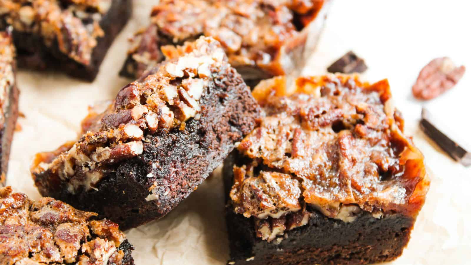Close-up of several chocolate brownies topped with pecans on a parchment surface.