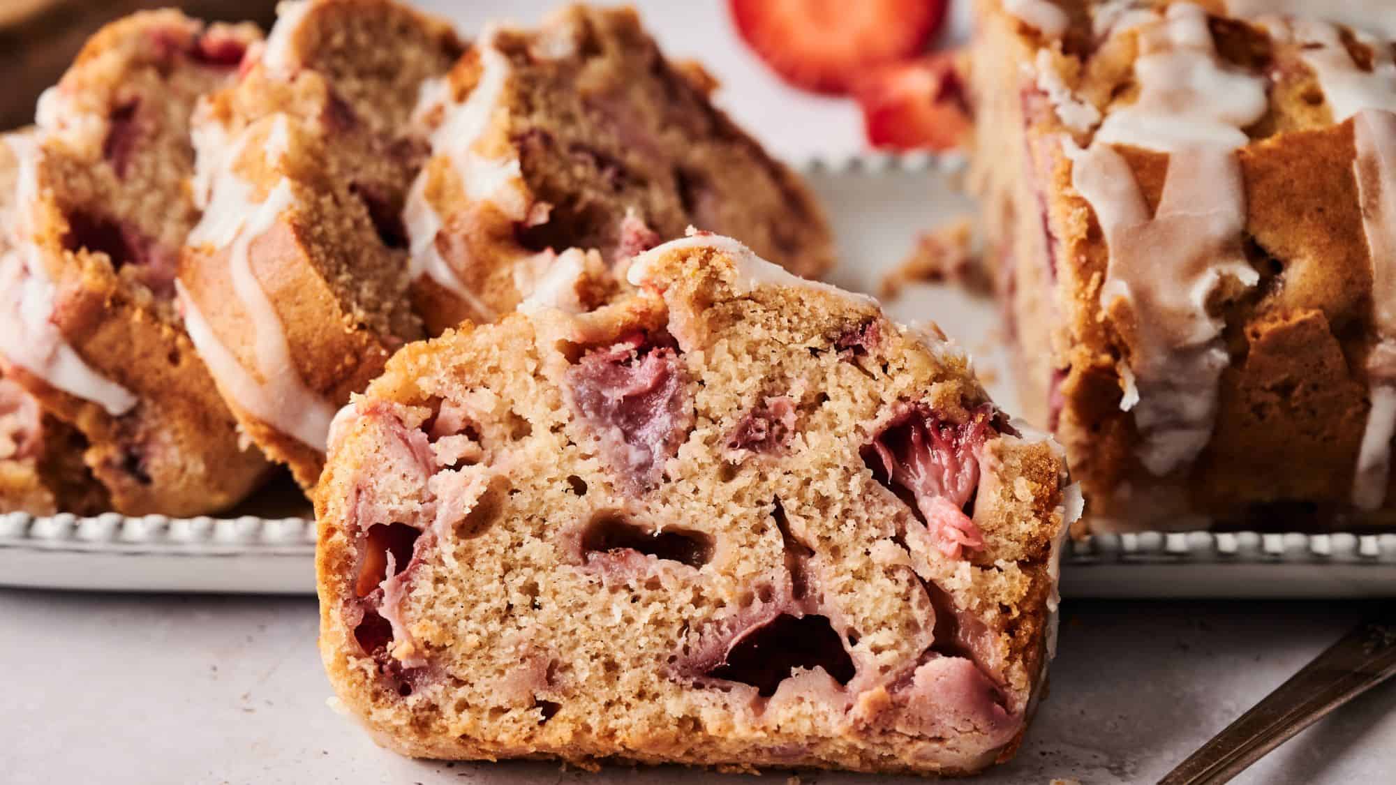 A close-up of sliced strawberry bread with visible strawberry pieces and a drizzle of white icing on top, arranged on a plate.