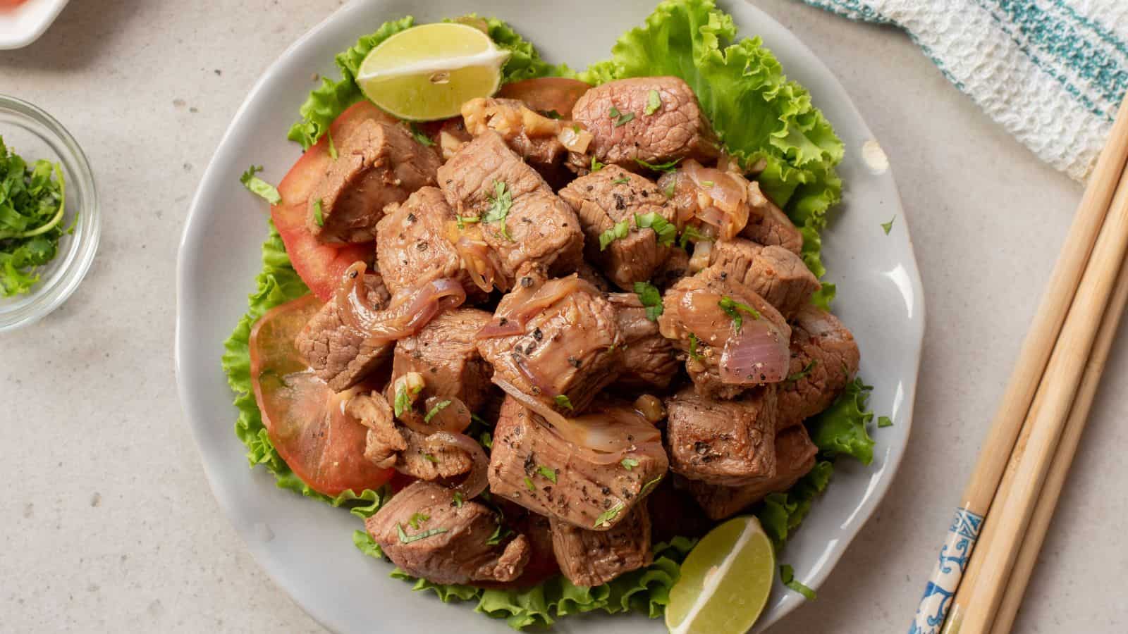 A plate of stir-fried beef cubes with onions, garnished with cilantro, served on lettuce with tomato slices and lime wedges. Chopsticks rest beside the plate.