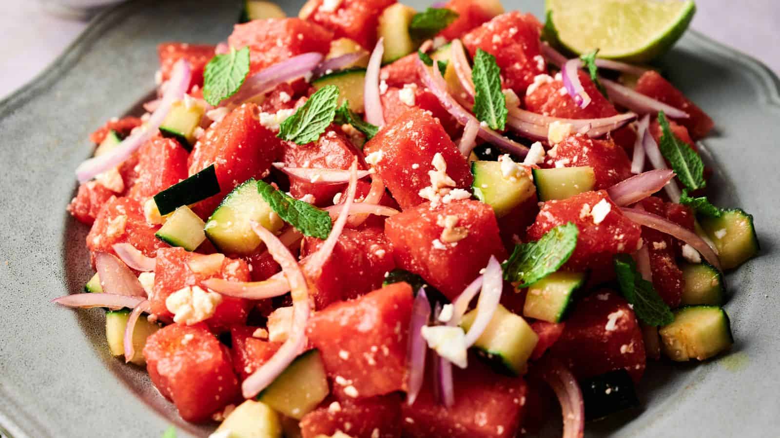 A plate of watermelon salad with cucumber, red onion, feta cheese, and mint leaves, served with a lime wedge.