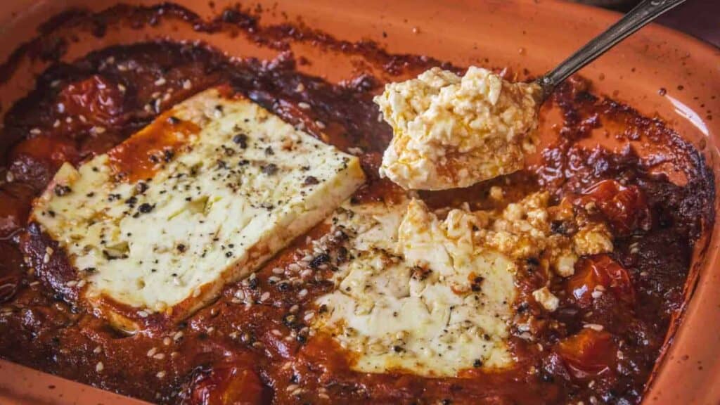 Baked feta cheese with roasted cherry tomatoes in a rectangular dish; a spoon lifts a portion of the cheese and tomato mixture.