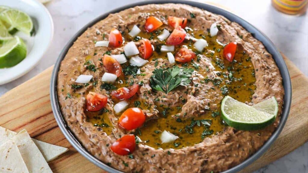 A bowl of refried beans garnished with chopped tomatoes, onions, cilantro, a lime wedge, and a drizzle of oil, served on a wooden board.
