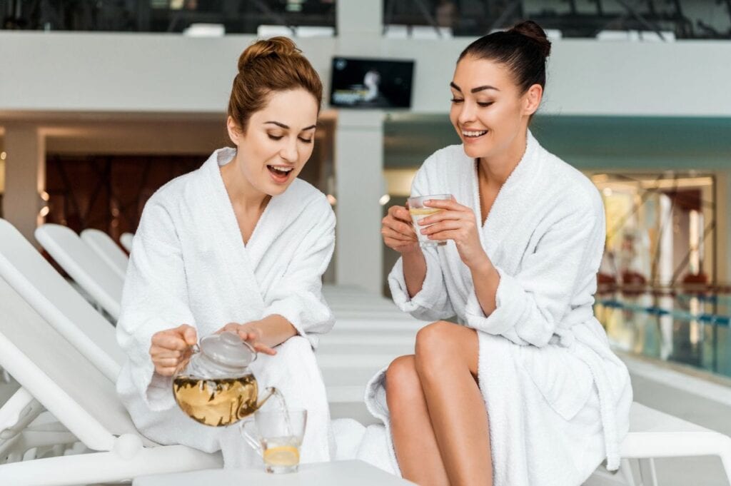 Two women in white robes sit by an indoor pool, one pouring tea while the other holds a glass, both relaxed and engaged in conversation&mdash;an ideal scene for summer sabbaticals.