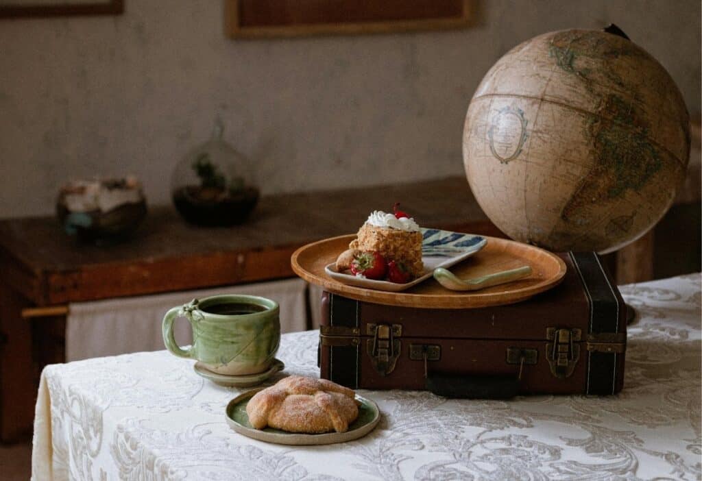 A slice of cake, a pastry, and a cup of coffee sit on a suitcase and table with a globe in the background.