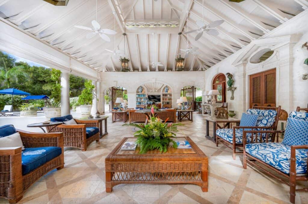 Spacious open-air living area with wicker furniture, blue cushions, potted plants, ceiling fans, and a view of a pool and garden&mdash;perfect for embracing the relaxed Barbados lifestyle.