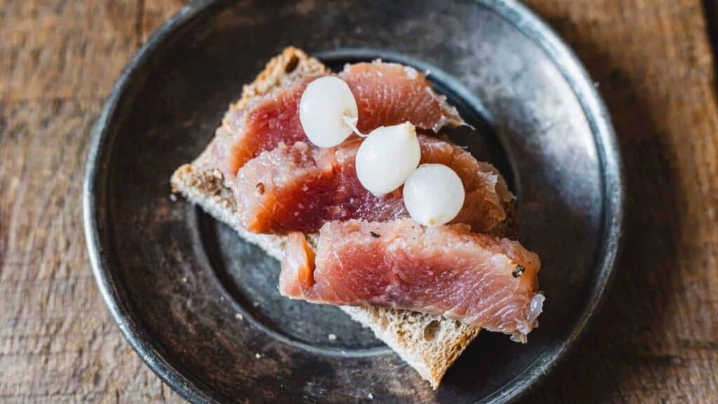 A slice of bread topped with pieces of raw or cured fish and three small pickled onions, served on a dark plate.