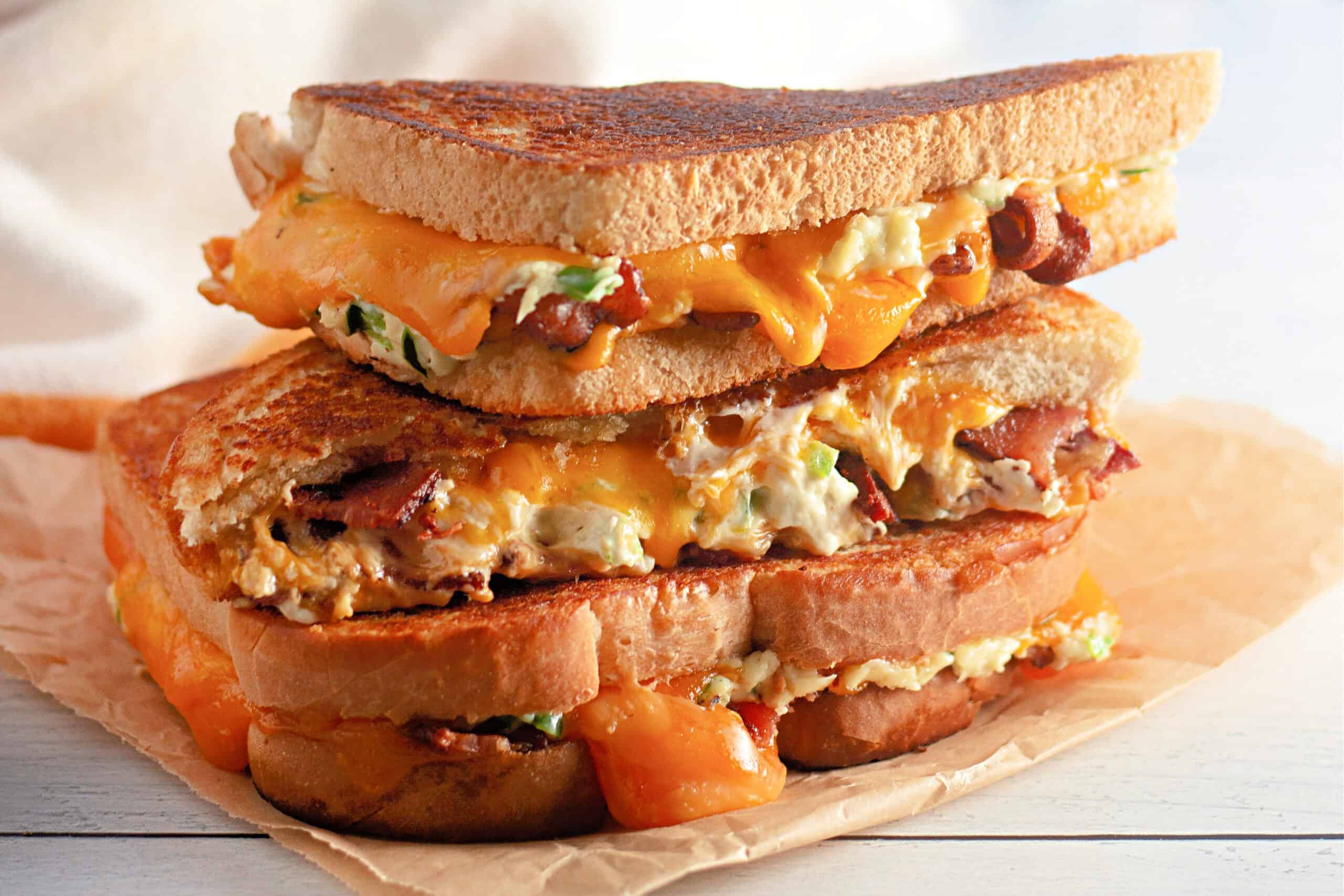 Close-up of two stacked grilled cheese sandwiches on parchment paper. Melted cheddar, bacon, and scallions ooze from crispy toasted bread, creating a warm, savory appeal.