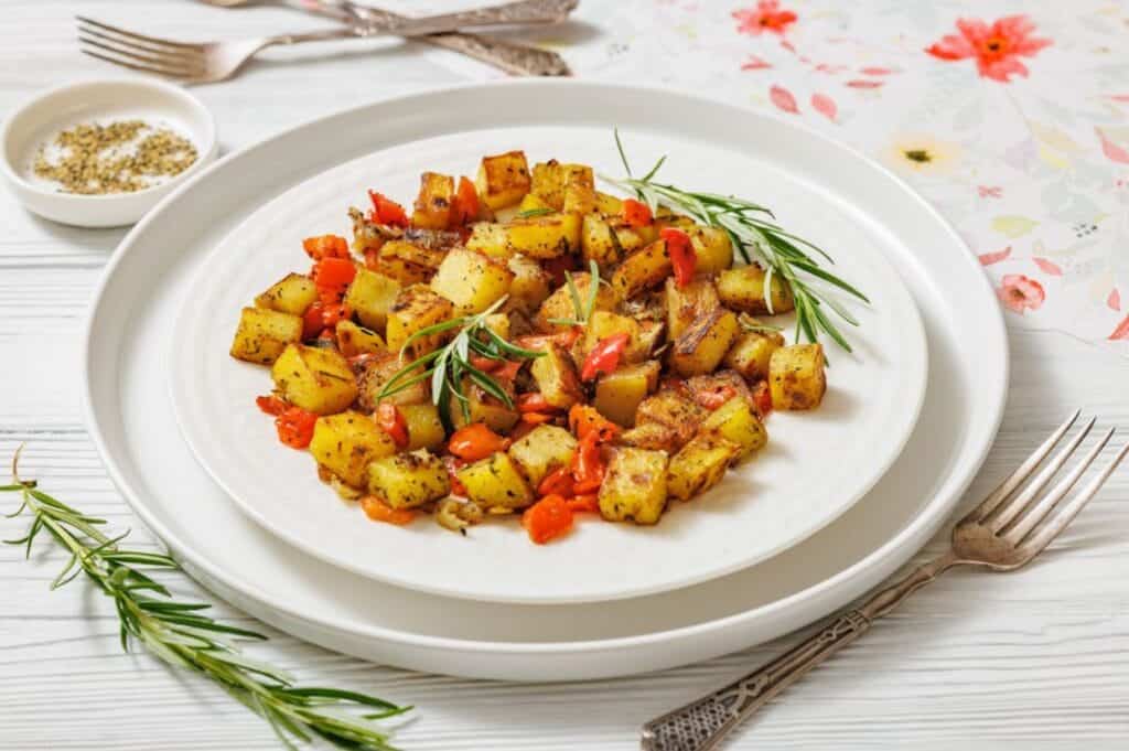 A white plate with Skillet Fried Potatoes garnished with rosemary sits on a white table with cutlery and a small bowl of pepper on the side.