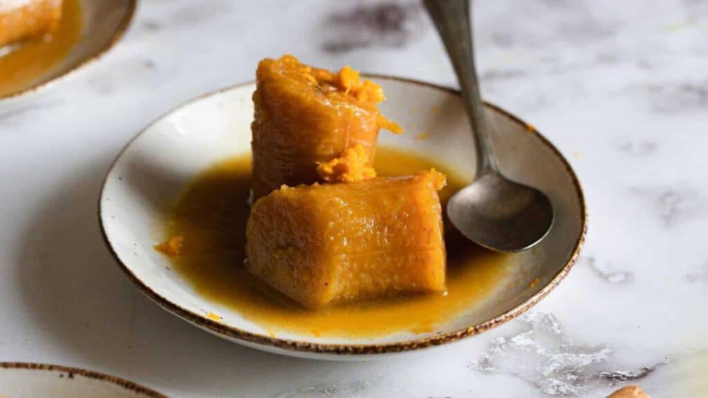 A small plate with two pieces of cooked plantain in syrup, with a spoon resting on the plate.