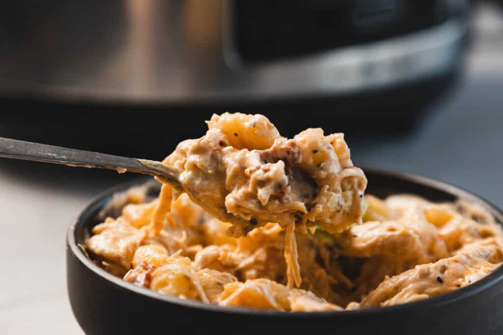 A close-up of a spoon lifting a serving of creamy, cheesy pasta from a black bowl, with a kitchen appliance in the background.