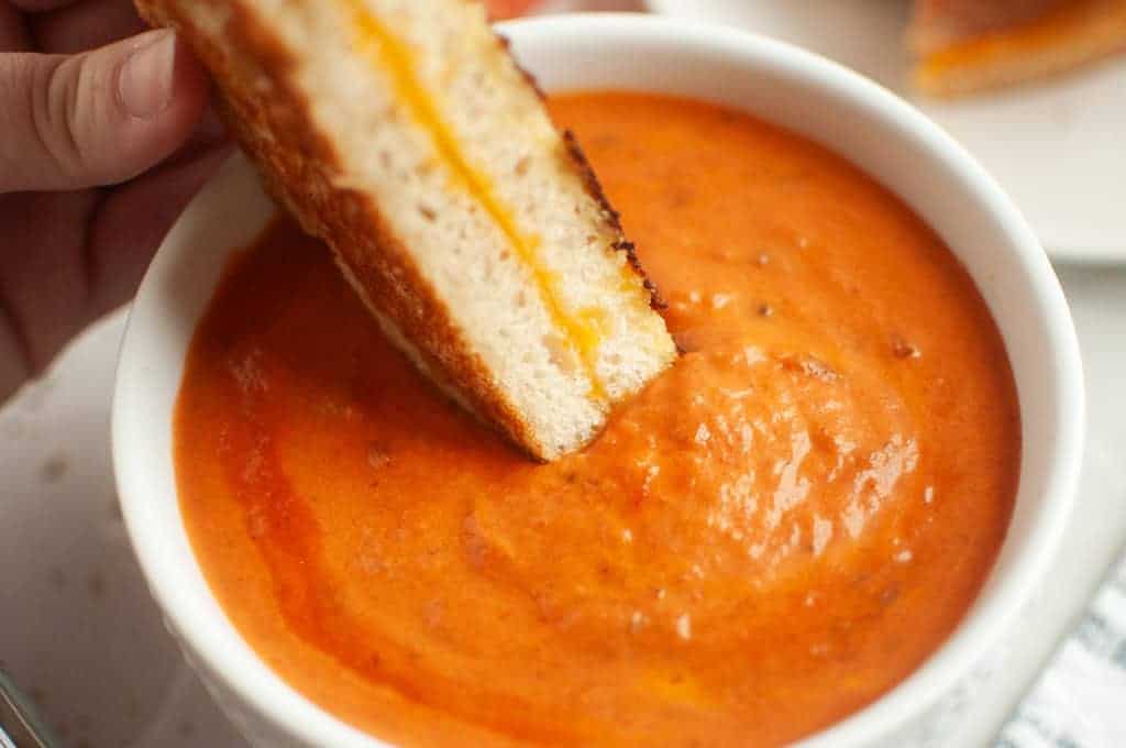 A hand dipping a piece of grilled cheese sandwich into a bowl of creamy tomato soup.