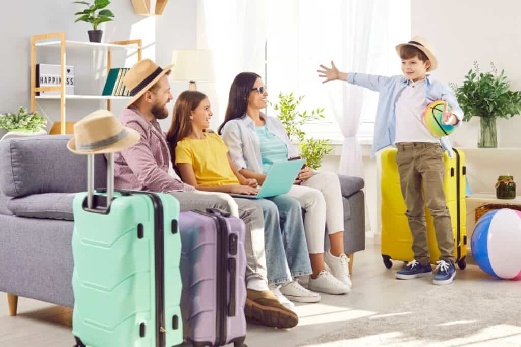 A family with suitcases sits on a couch while a child stands nearby holding a beach ball, as they gather their road trip essentials and prepare for an exciting vacation.